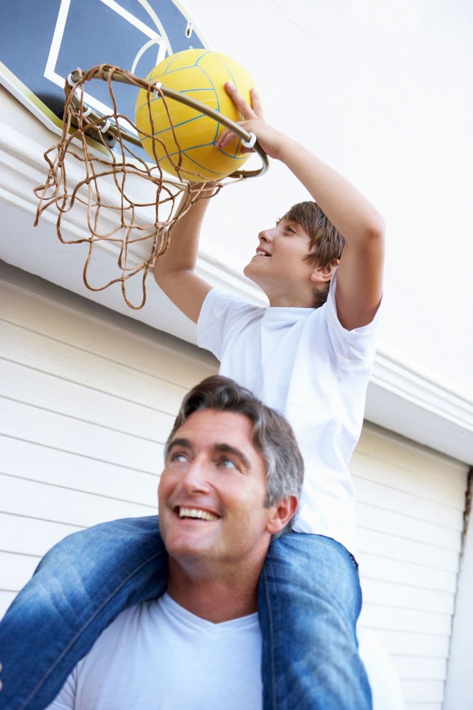 son on shoulders playing basketball in front of home garage