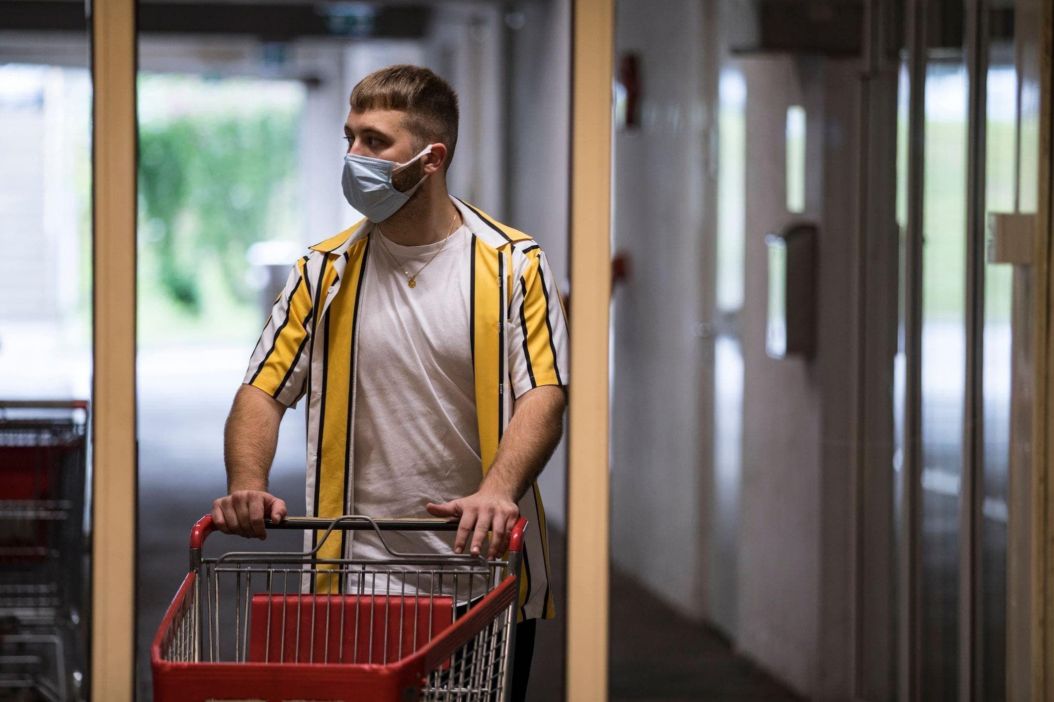 a young man wearing a medical face mask pushes a shopping cart to enter automatic grocery store doors